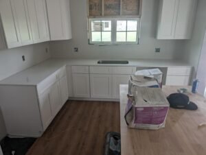 Newly installed white kitchen cabinets and countertops with an unfinished window and unpacked materials on the island, during a kitchen renovation project.