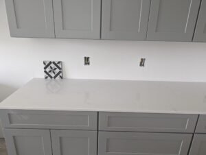 Gray kitchen cabinets with a white countertop and a patterned tile sample placed against the wall, part of an ongoing renovation project.